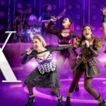 Six performers in colourful costumes sing and dance on a purple-lit stage in SIX The Musical at the Vaudeville Theatre London