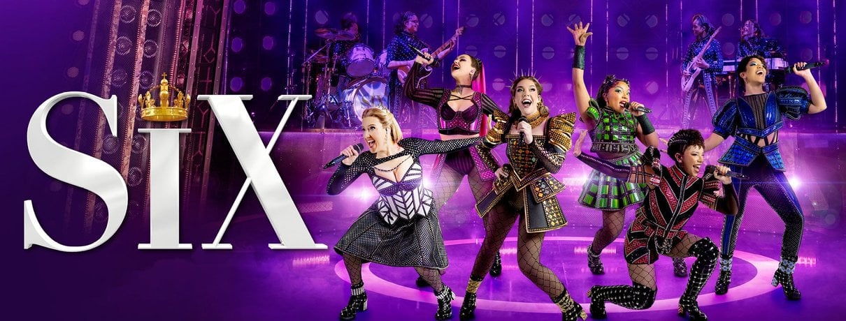 Six performers in colourful costumes sing and dance on a purple-lit stage in SIX The Musical at the Vaudeville Theatre London