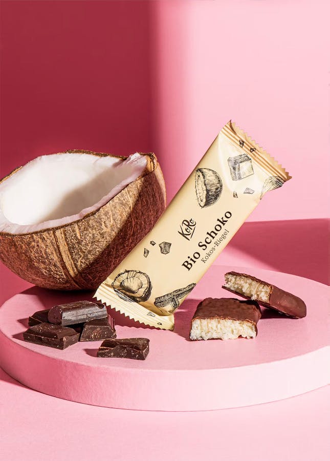 Bio Schoko-Kokos-Riegel neben einer Kokosnuss auf rosa Hintergrund
