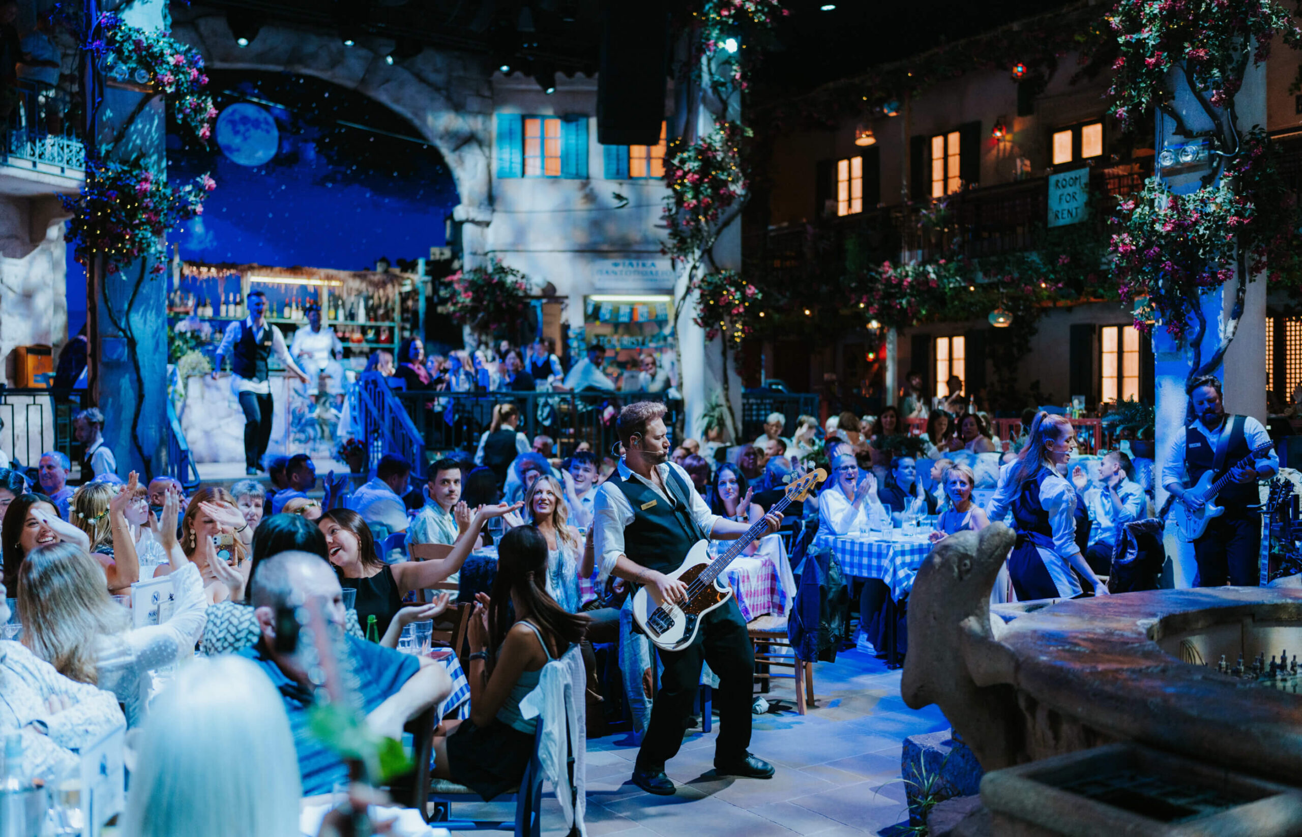Live band member playing bass among the diners under blue light at Mamma Mia! The Party