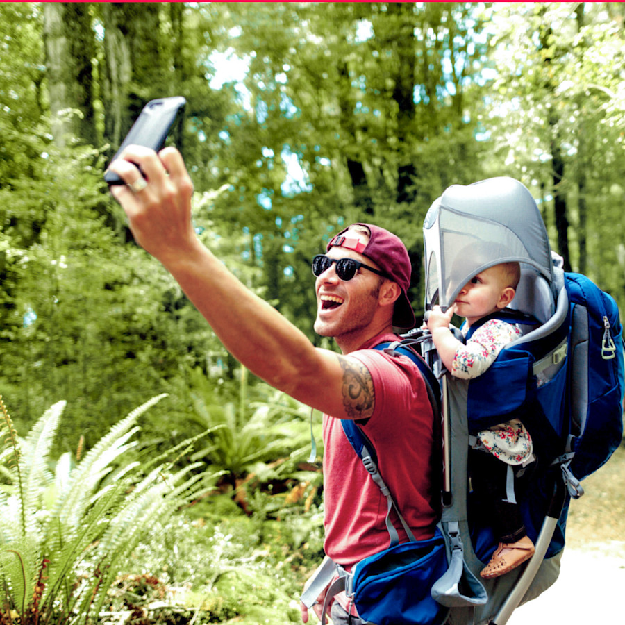Vater mit Kind in der Rückentrage macht beim Wandern ein Selfie mit dem Smartphone