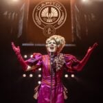 A performer as Effie Trinket singing beneath the Capitol emblem on stage
