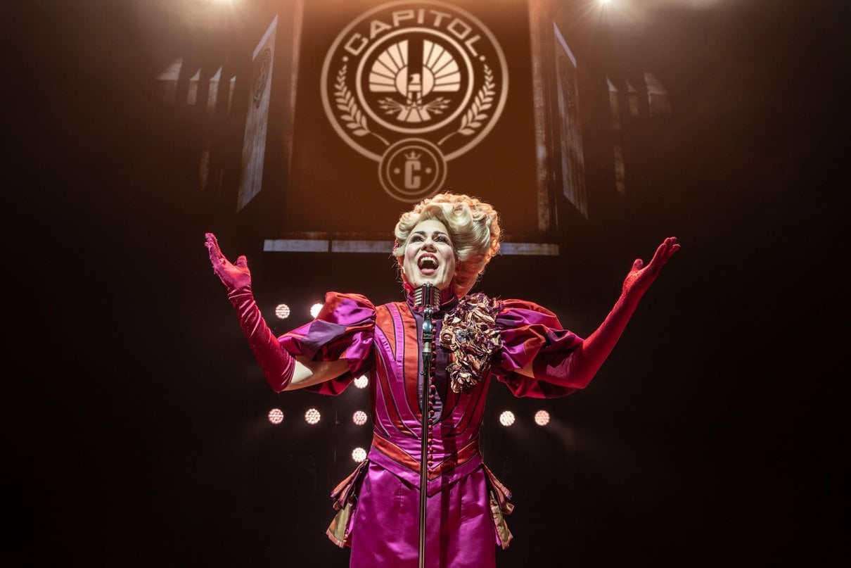 A performer as Effie Trinket singing beneath the Capitol emblem on stage