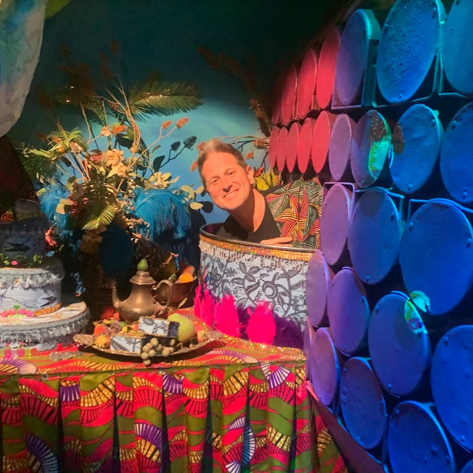 Visitor behind a colourful tin wall with blue barrels at JAMNOLA